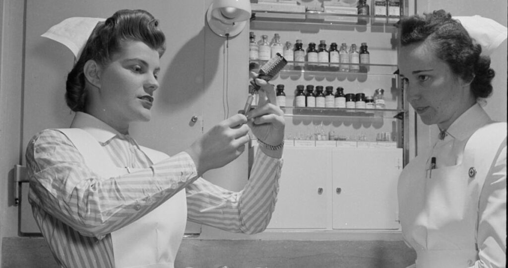 Black and white photo of two women wearing nurses uniforms in what appears to be a medical office. One is drawing liquid from a vial into a syringe as the other watches.